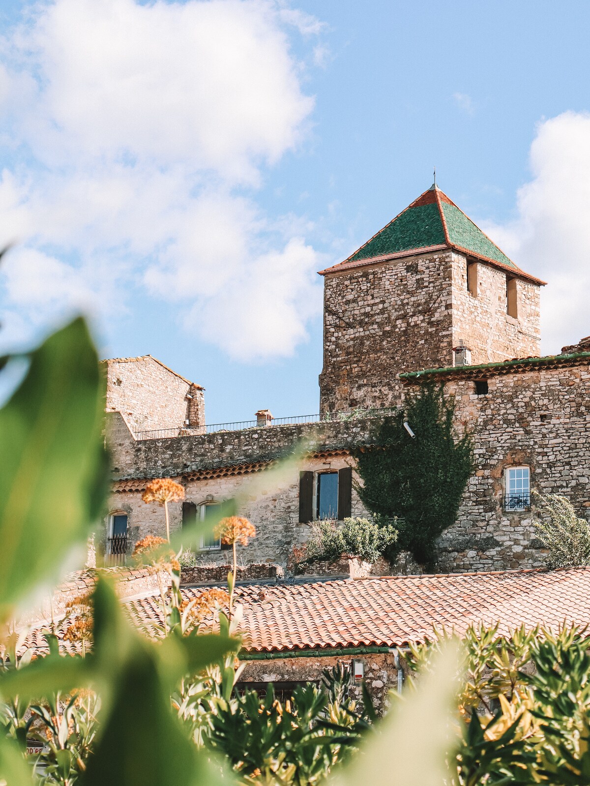 A stone tower with a pointed green roof is seen rising from a historic building. Lush green plants and flowers frame the bottom of the image, offering a glimpse of rustic architecture under a blue sky with scattered clouds.