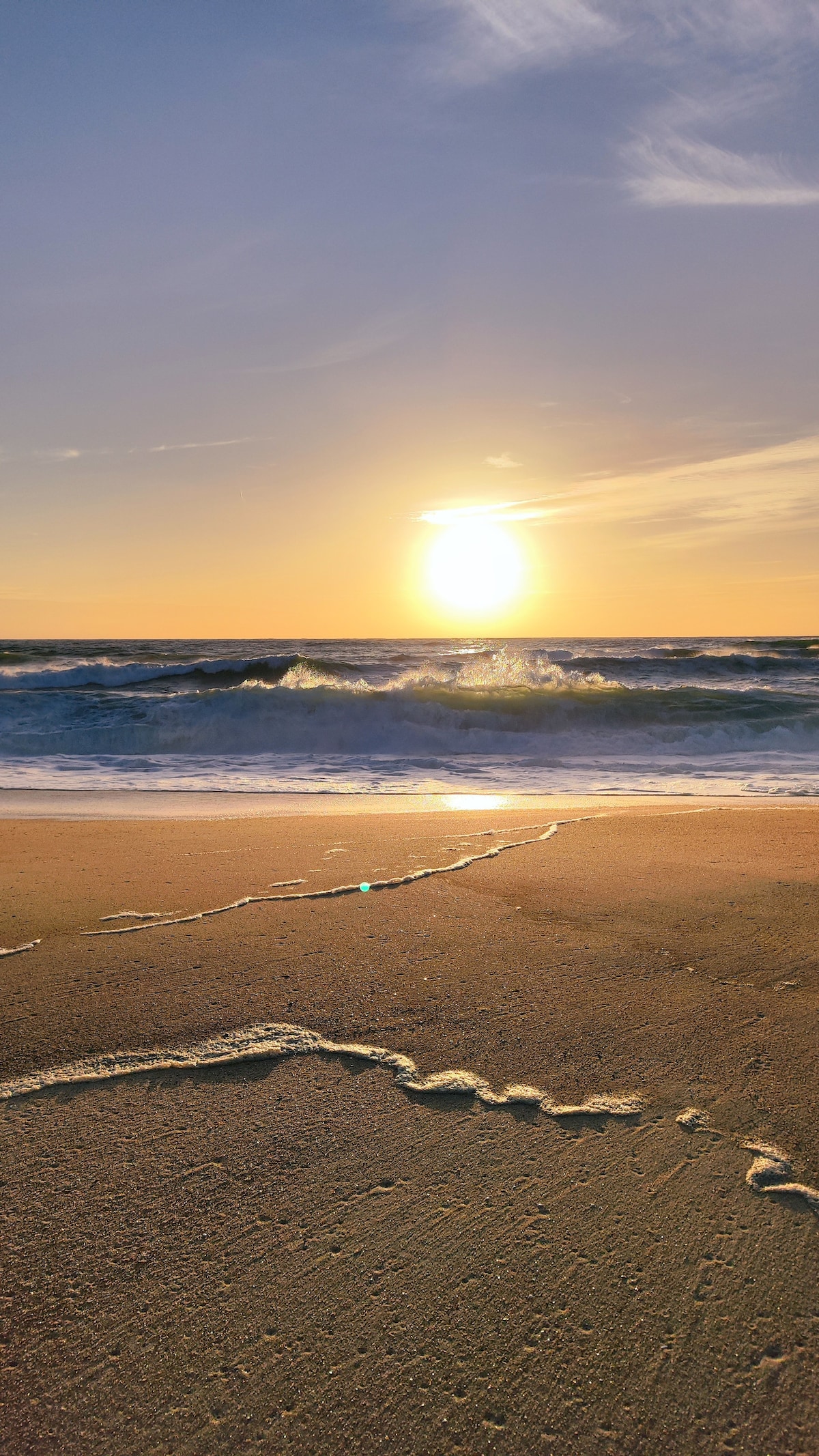 The serene beach scene captures sunlight reflecting on gentle waves as they roll onto the shore. The sand is marked with subtle patterns from water receding. A warm golden hue envelops the landscape, indicating the time of sunset, creating a calming atmosphere.