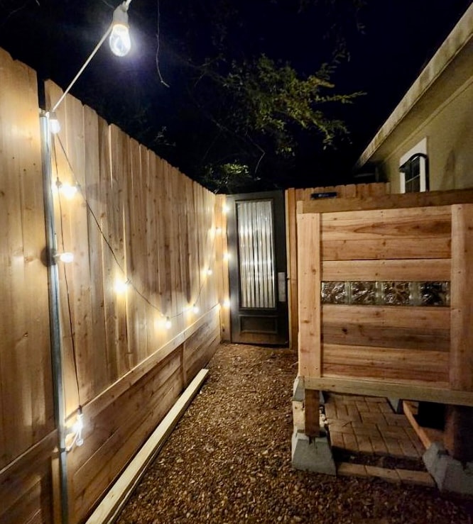 A narrow pathway is illuminated by warm string lights, creating a welcoming atmosphere at night. A wooden fence borders the path, while a glass-paneled door stands at the end, inviting further exploration.