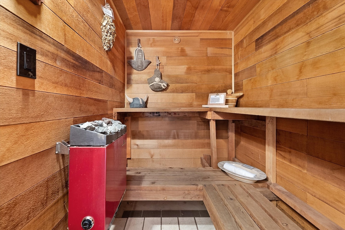 A calming sauna space features warm wooden walls and benches, enhancing the natural aesthetic. A red heater sits against one wall, and various decorative items, including a wooden paddle and tools, are displayed around the area, contributing to the serene environment.