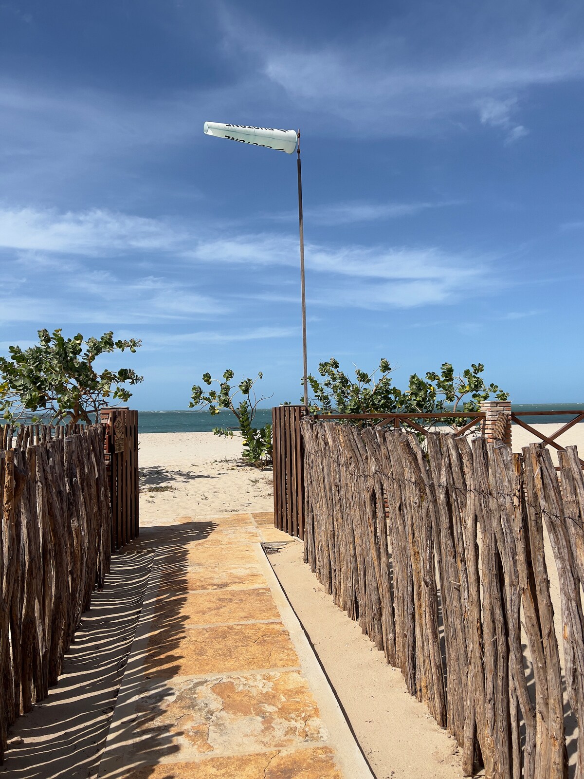 A pathway lined with rustic wooden fencing leads to the sandy beach. The bright blue sky and calm sea are visible in the background, creating a serene coastal setting. Nearby greenery adds a natural touch to this inviting entrance to the shore.