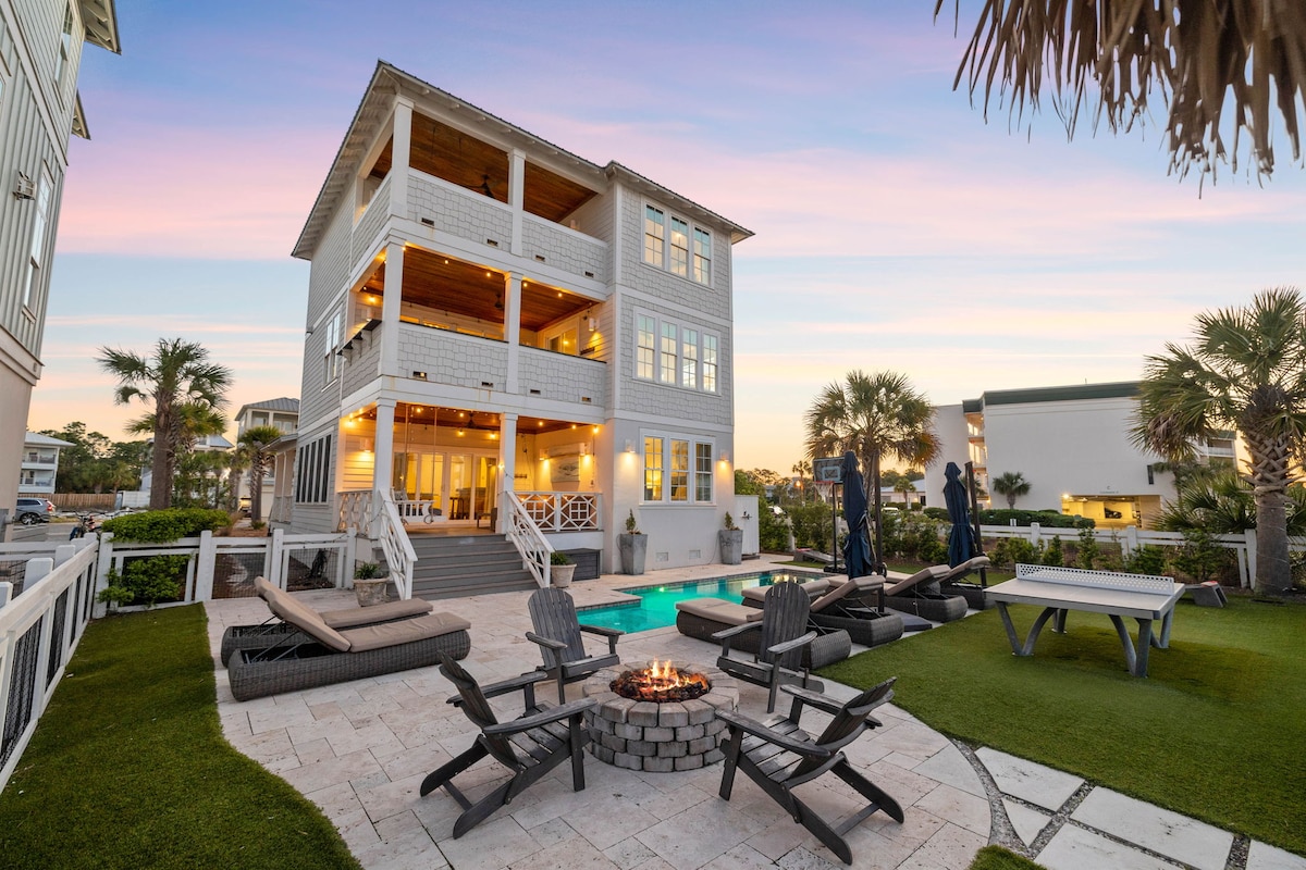 The exterior of the three-story home features a spacious backyard with a private saltwater pool and a fire pit surrounded by adirondack chairs. Palm trees frame the scene, while multiple seating areas provide space for relaxation and enjoyment, all set against a colorful sunset sky.