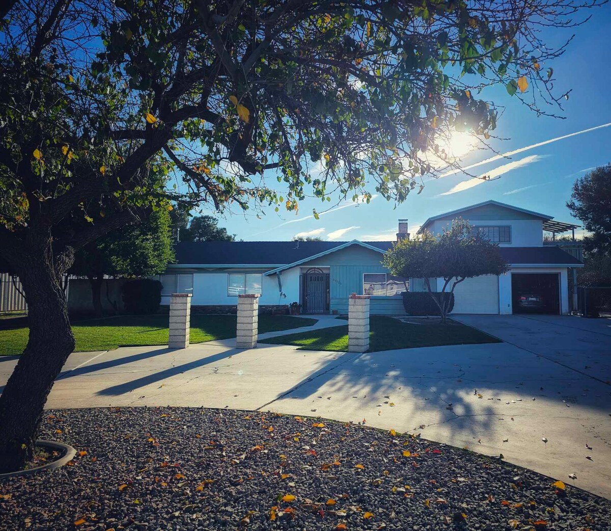 A single-story house is framed by a lush green lawn and mature trees. Sunlight shines on the home, casting gentle shadows on the concrete driveway. Decorative stone pillars line the entrance, contributing to the welcoming atmosphere of the property.