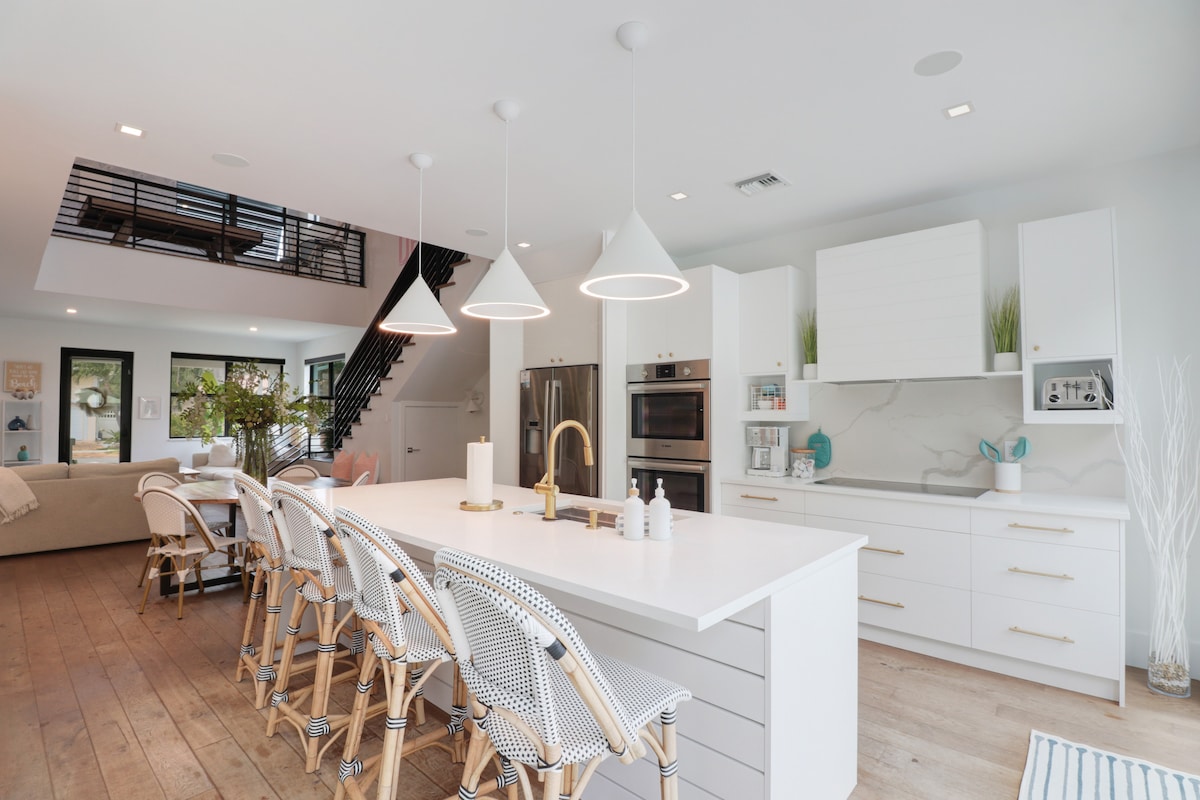 A modern kitchen is equipped with white cabinetry and stainless steel appliances. A spacious island features seating for several, complemented by stylish bar stools. Natural light pours in through large windows, highlighting the open floor plan that connects to the living space.