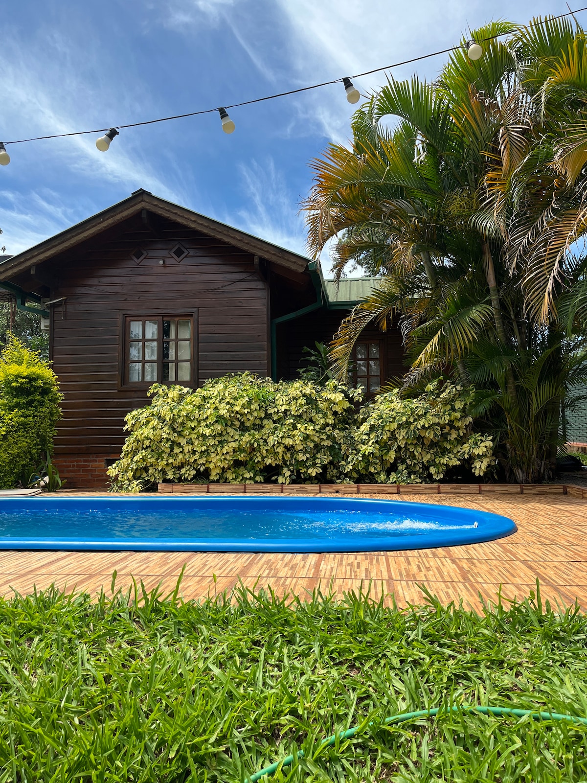 A wooden cabin is set against a backdrop of lush greenery and blooming shrubs. A rectangular pool is bordered by a bamboo deck, inviting relaxation. String lights are visible above, adding a gentle point of interest to the outdoor space.