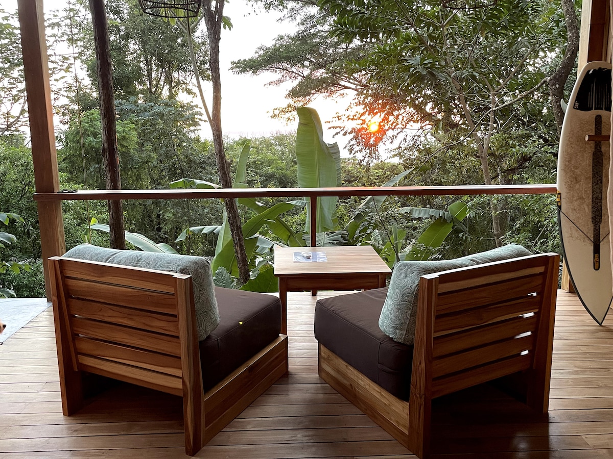 Two wooden armchairs with thick cushions face a small wooden table, positioned on a spacious deck. Lush greenery surrounds the area, while the sun sets in the background, casting a warm glow. The scene offers a serene outdoor setting, perfect for relaxation.