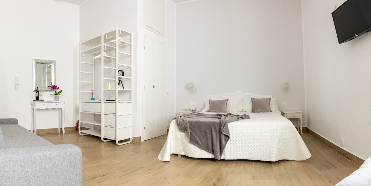A spacious bedroom features a large bed with soft linens, flanked by wall sconces. A light-colored wardrobe and a sleek white mirror add functionality. A small table is positioned beside the bed, while a gray couch occupies one corner, ensuring a comfortable and inviting setting.