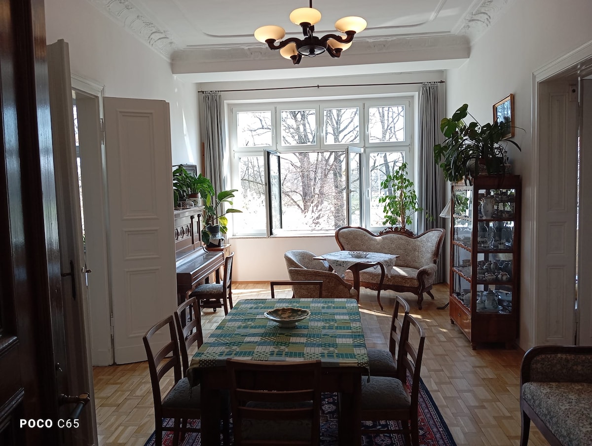 A dining area features a wooden table surrounded by chairs, with a patterned tablecloth at the center. Natural light floods through large windows, illuminating a cozy seating arrangement. Potted plants add greenery, and a cabinet displays various items along one wall.