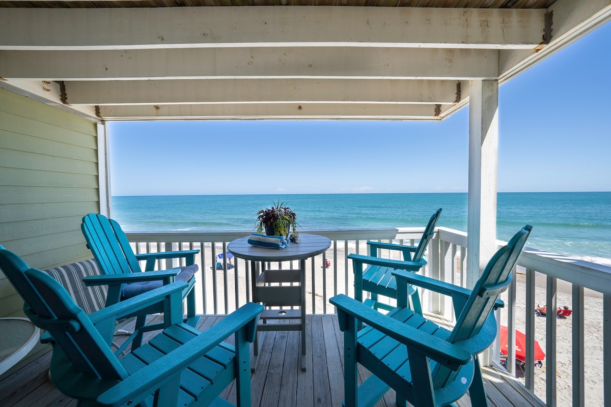 A spacious balcony features four blue Adirondack chairs arranged around a small table. The ocean view is unobstructed, with waves gently lapping on the shore. Sunlight illuminates the scene, creating a calm and inviting outdoor space.