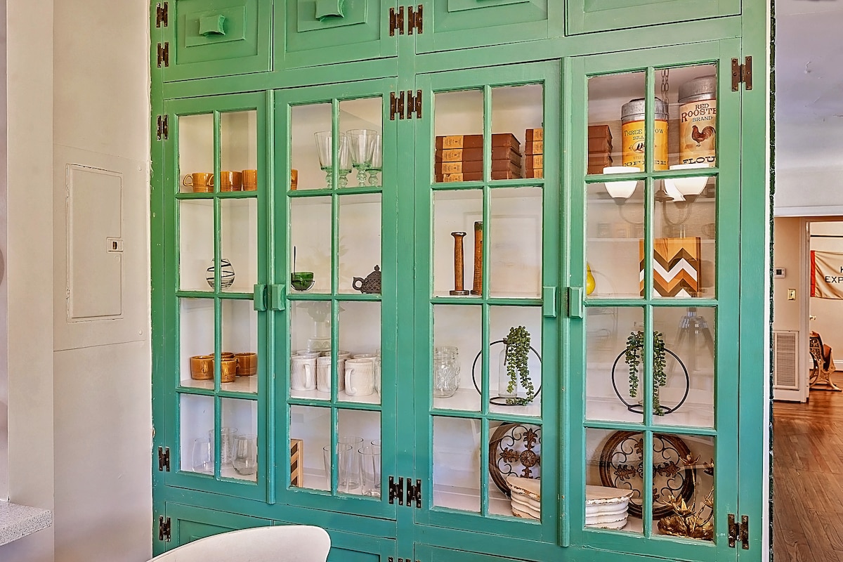 A vibrant green cabinet with glass doors showcases an array of decorative items. Inside, various containers, glasses, and stylish decor pieces are neatly arranged, contributing to a welcoming interior ambiance. Soft light illuminates the contents, enhancing the visual appeal of the displayed items.