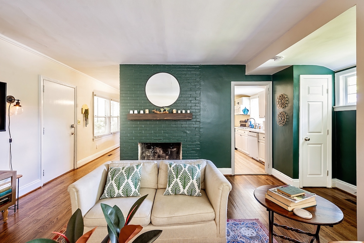 A comfortable living area features a light-colored sofa adorned with patterned cushions. A green accent wall houses a round mirror above a decorative shelf. Natural light enters through windows, illuminating the space, which flows into a well-lit kitchen area visible through an open doorway.