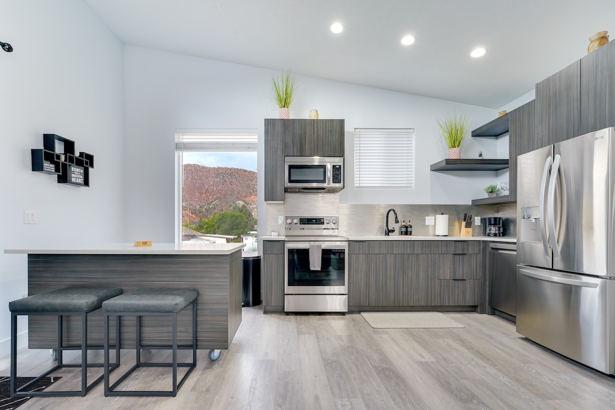 A modern kitchen showcases sleek cabinetry and stainless steel appliances, complemented by a spacious island with two bar stools. Natural light pours through a window, revealing views of the surrounding red mountains. A neutral color palette enhances the contemporary design.