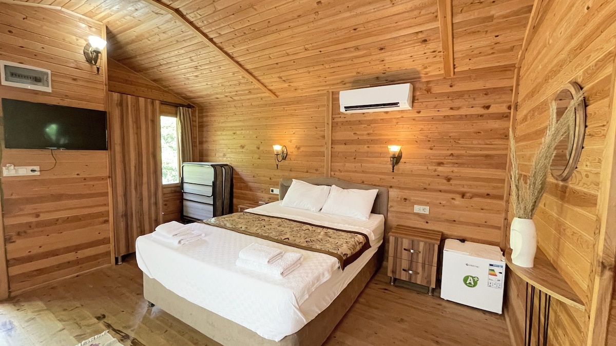 A wooden cabin interior features warm wooden paneling and a spacious bed with crisp linens. Two wall sconces provide soft lighting, and a television is mounted on the wall. A compact refrigerator is positioned beside a storage unit, and a window allows natural light to enter.