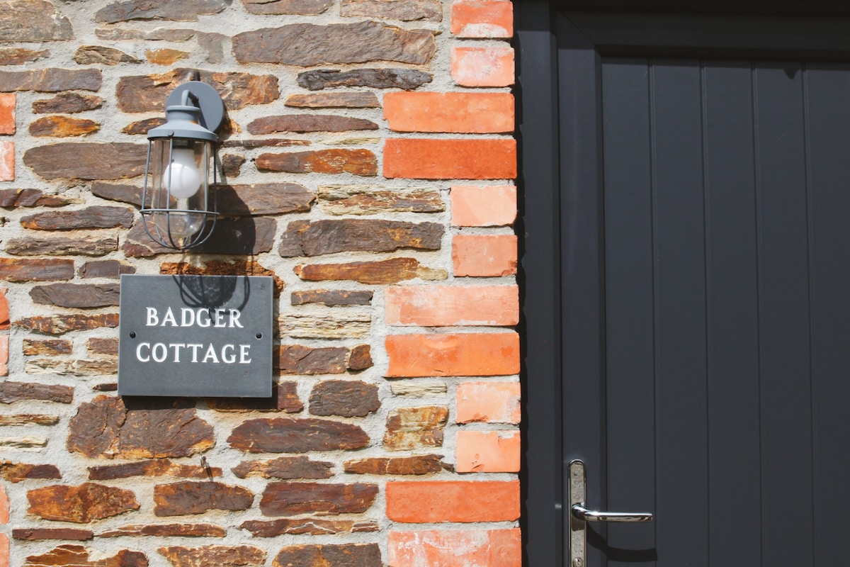 The exterior of Badger Cottage is framed by a mix of rustic stone and warm red bricks. A subtle light fixture is mounted beside a solid black door adorned with a sleek handle. A nameplate with 'Badger Cottage' is prominently displayed.