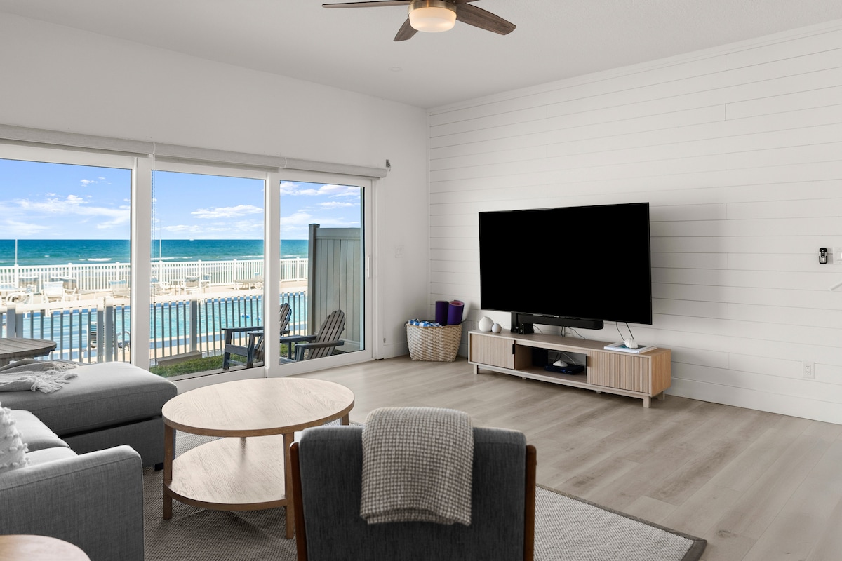 A bright living area features large glass doors that open to reveal views of the pool and ocean. A spacious sectional sofa is accompanied by a round coffee table. A flat-screen TV is placed on a light wood media console, against a backdrop of shiplap walls.