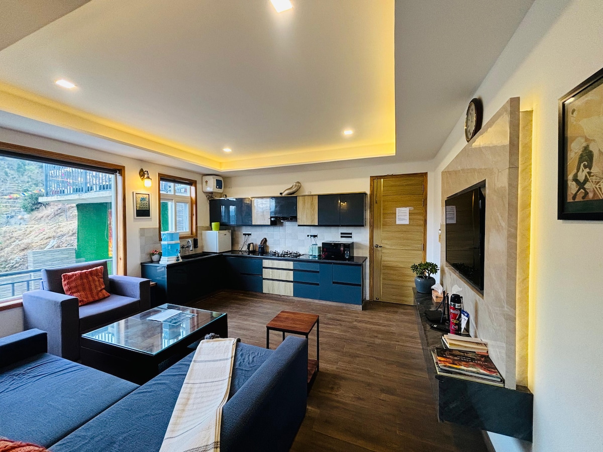 A modern living area features a spacious sofa, a flat screen TV, and large windows allowing natural light. The kitchen is equipped with contemporary appliances and cabinetry. A dining table for four is visible, and a decorative wall unit adds character to the space.