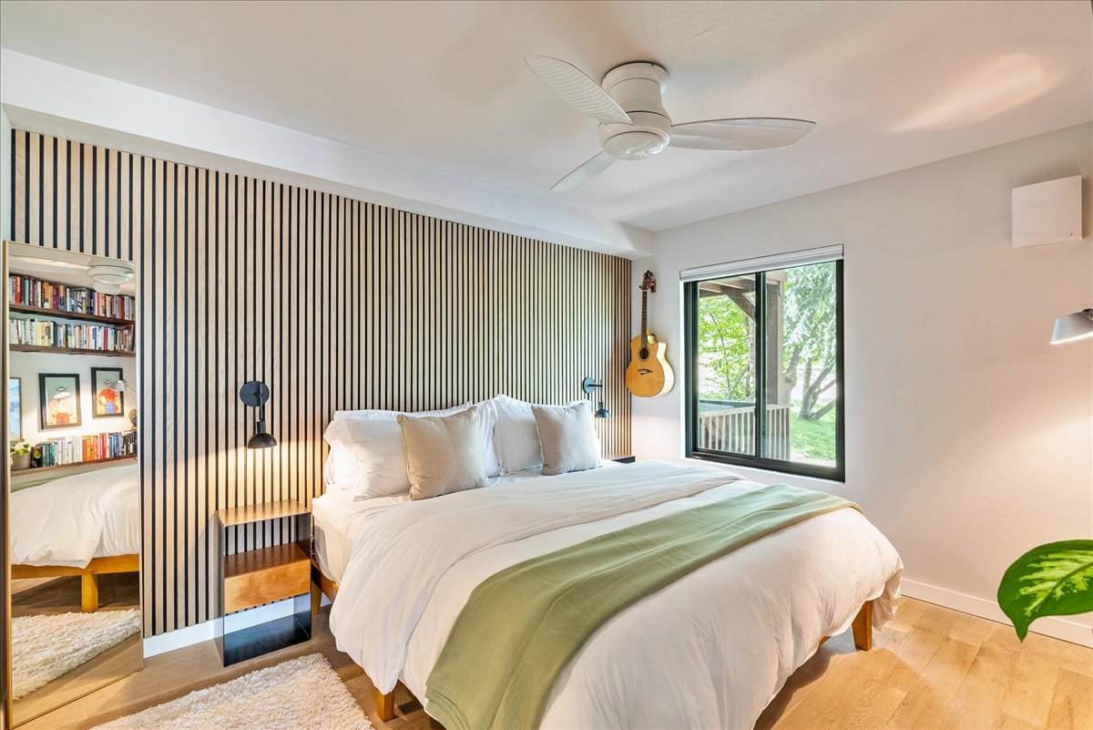A bright bedroom features a spacious bed with layered bedding and decorative cushions. A wooden accent wall enhances the room's design. Natural light streams through the window, offering a view of greenery. A guitar and a small lamp are present, adding character to the space.