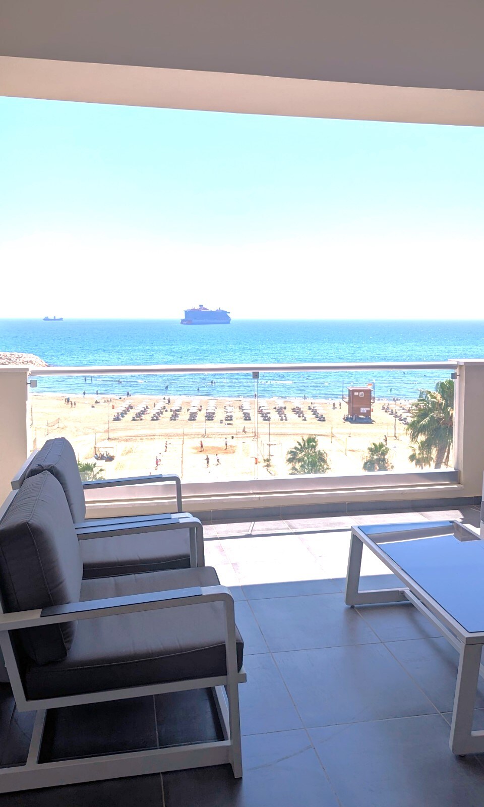 A covered balcony features modern outdoor furniture, including a sofa and table, overlooking a serene view of the Mediterranean Sea. The scene includes a sandy beach with organized sun loungers and umbrellas, creating a tranquil coastal atmosphere.
