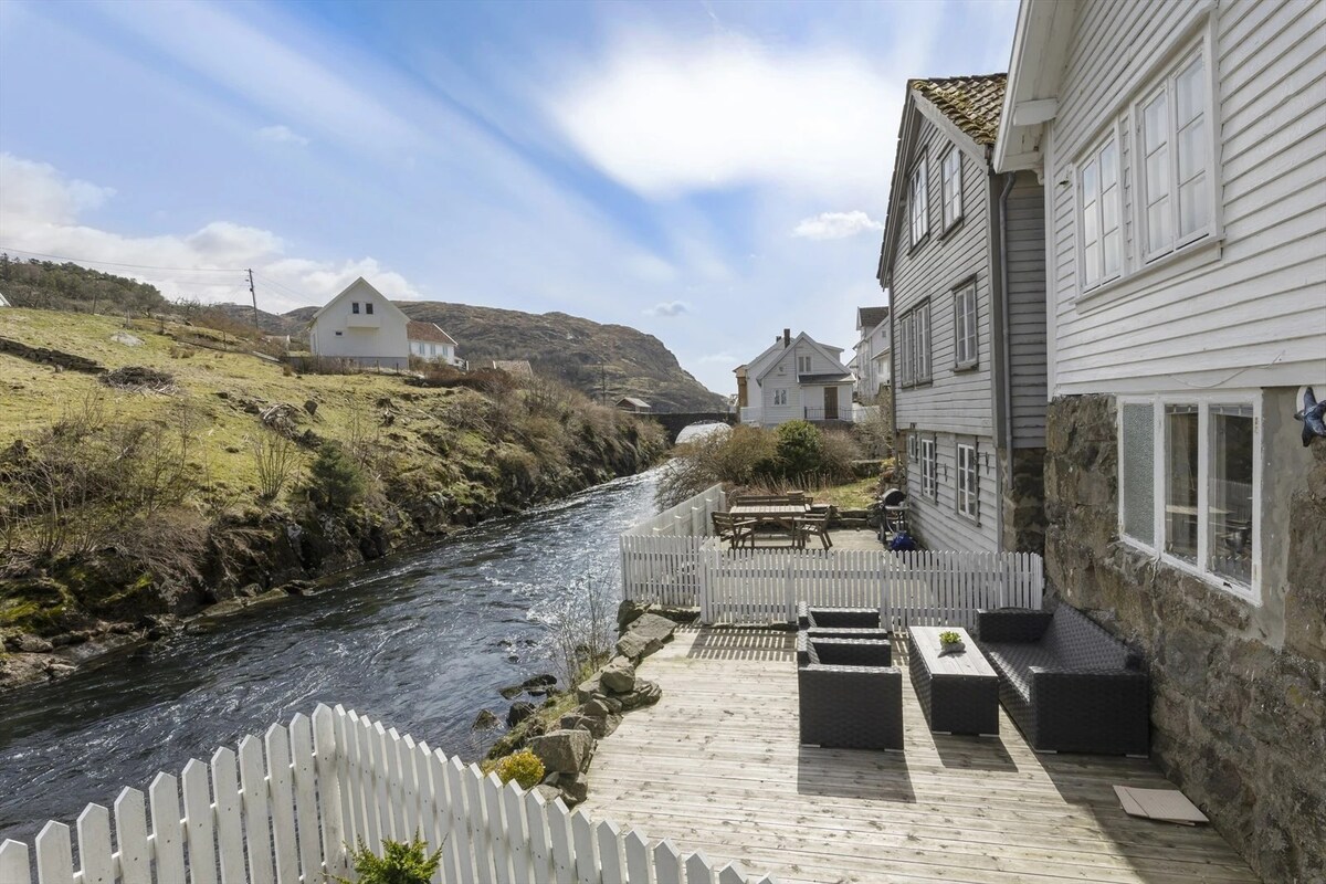 A spacious patio features comfortable seating and a dining area, overlooking the flowing river. The wooden deck is bordered by a white picket fence. Lush green hills rise in the background, with charming houses in the distance under a clear blue sky.