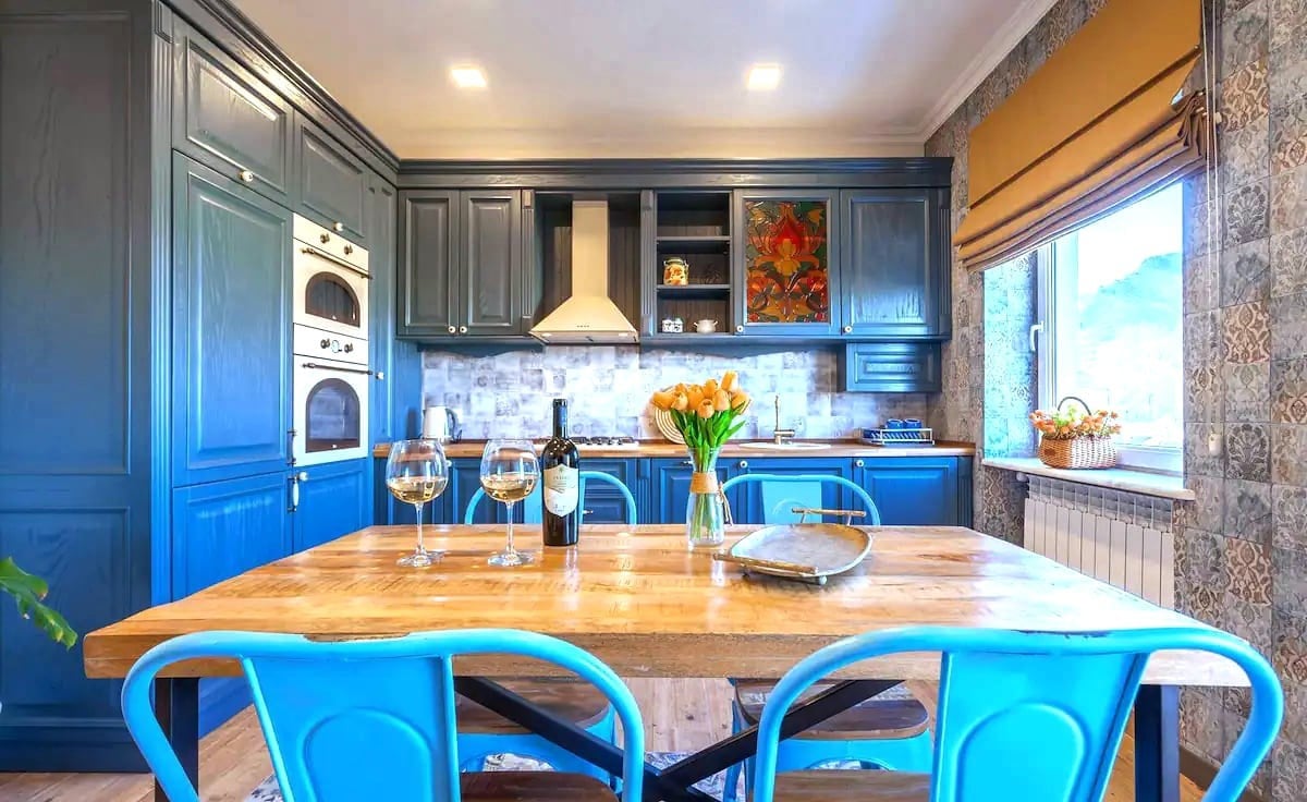 A modern kitchen area is present, featuring rich blue cabinetry and a wooden dining table set for four. Glassware sits beside a bottle of wine, and a vase of tulips adds color. Natural light comes through a window with soft window treatments.