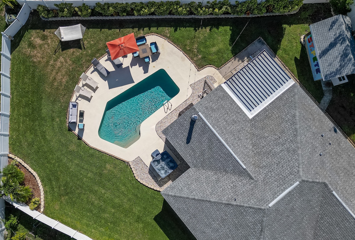 An aerial view showcases a spacious backyard with a sparkling pool at its center, surrounded by sun loungers and an orange umbrella. The patio features various outdoor furniture arrangements, while the well-maintained lawn adds to the inviting atmosphere.