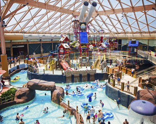 An expansive indoor waterpark features various water slides, play structures, and pools. Families can be seen enjoying the attractions, with tubes floating along a lazy river. A bright, transparent roof allows natural light to fill the space, enhancing the lively atmosphere.