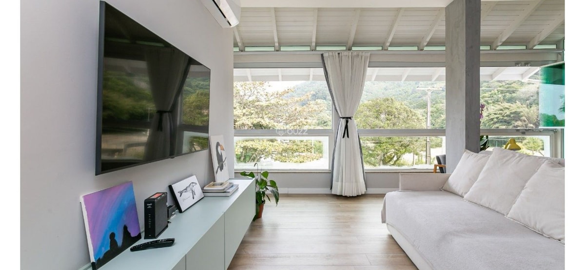 A modern living area is illuminated by natural light from large floor-to-ceiling windows. A sleek television is mounted on the wall, with a minimalist console beneath it. A light-colored sofa is positioned nearby, accompanied by a small indoor plant and gentle drapes framing the view.