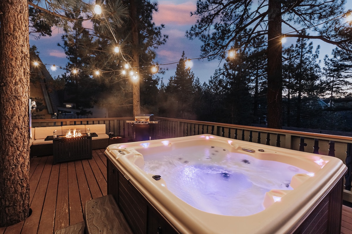 A hot tub is centered on a wooden deck, illuminated by soft café lights above. Surrounding trees provide a natural backdrop, while a firepit is situated nearby, creating a cozy atmosphere for relaxation. The evening sky features gentle hues as twilight approaches.