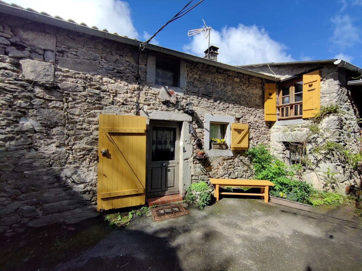 A rustic stone cottage exterior features yellow wooden shutters and a welcoming front door. Lush greenery surrounds the entrance, with a yellow bench positioned nearby. A small window above the door adds character, while the sky is bright and partly cloudy.