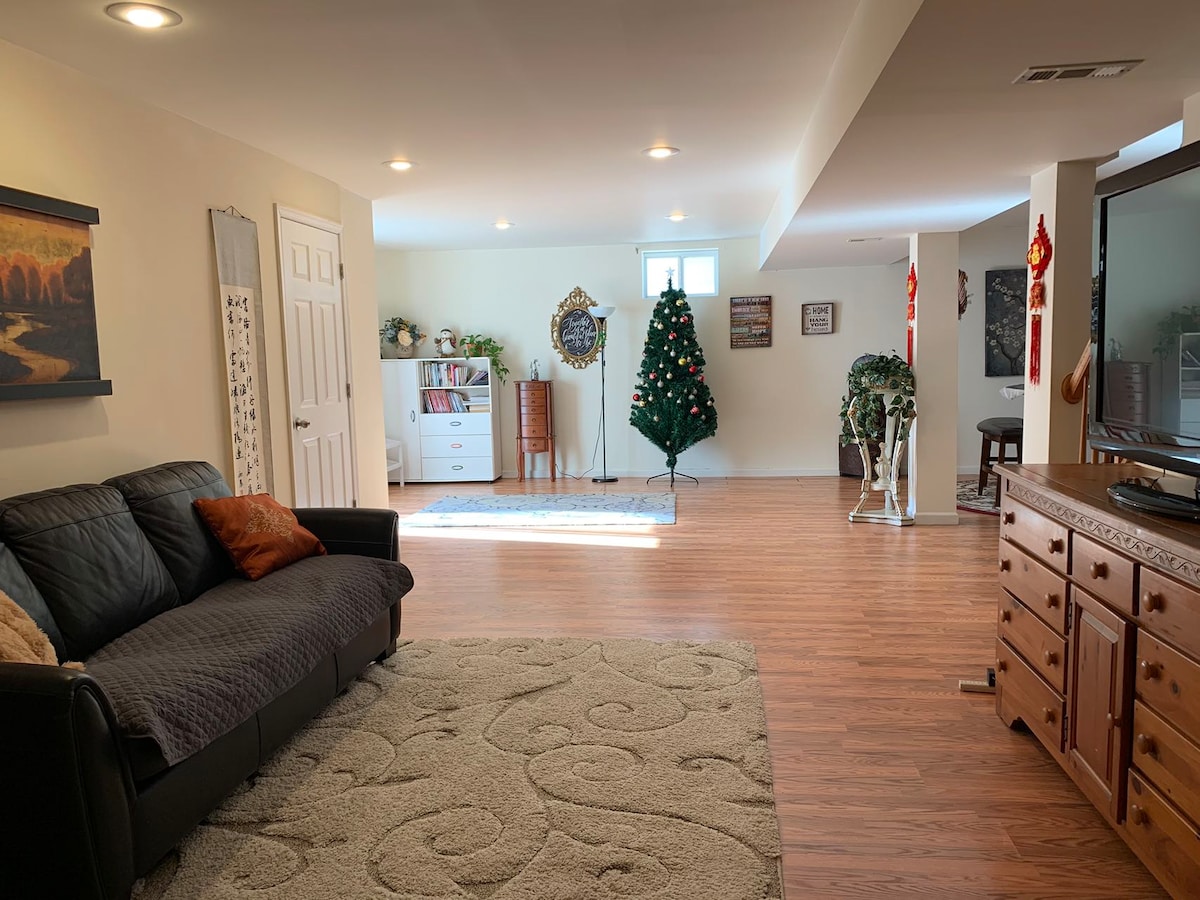 A spacious living room is highlighted by warm wood flooring and soft overhead lighting. A cozy sofa, accompanied by decorative pillows, faces a media unit. A large artificial Christmas tree stands in the corner, with a few decorative items visible on a nearby shelf.