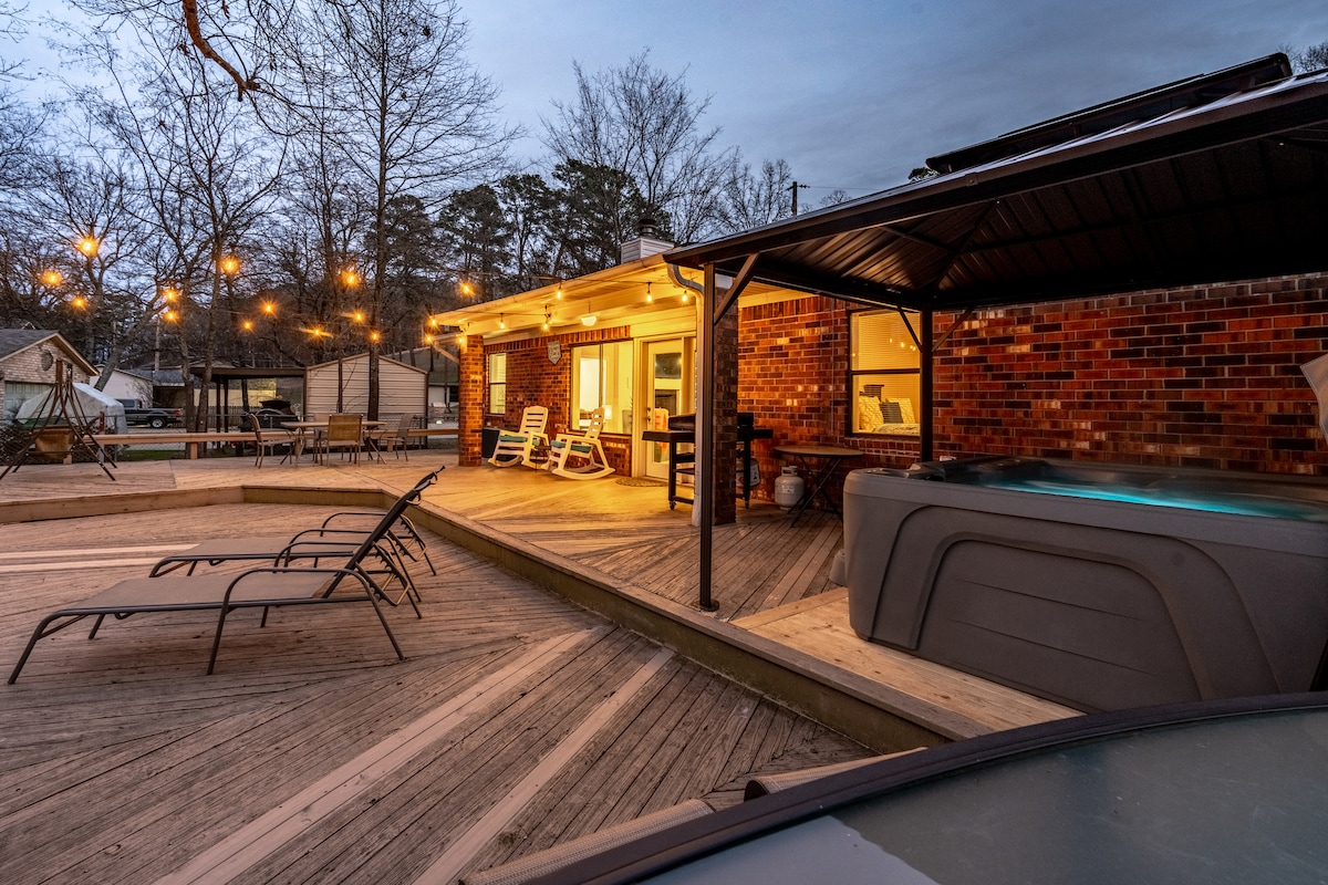 A spacious outdoor deck is illuminated by soft string lights, creating a warm ambiance. A hot tub is positioned nearby, alongside comfortable lounge chairs and a couple of white rocking chairs. The rustic brick home is visible in the background, with windows reflecting the evening light.