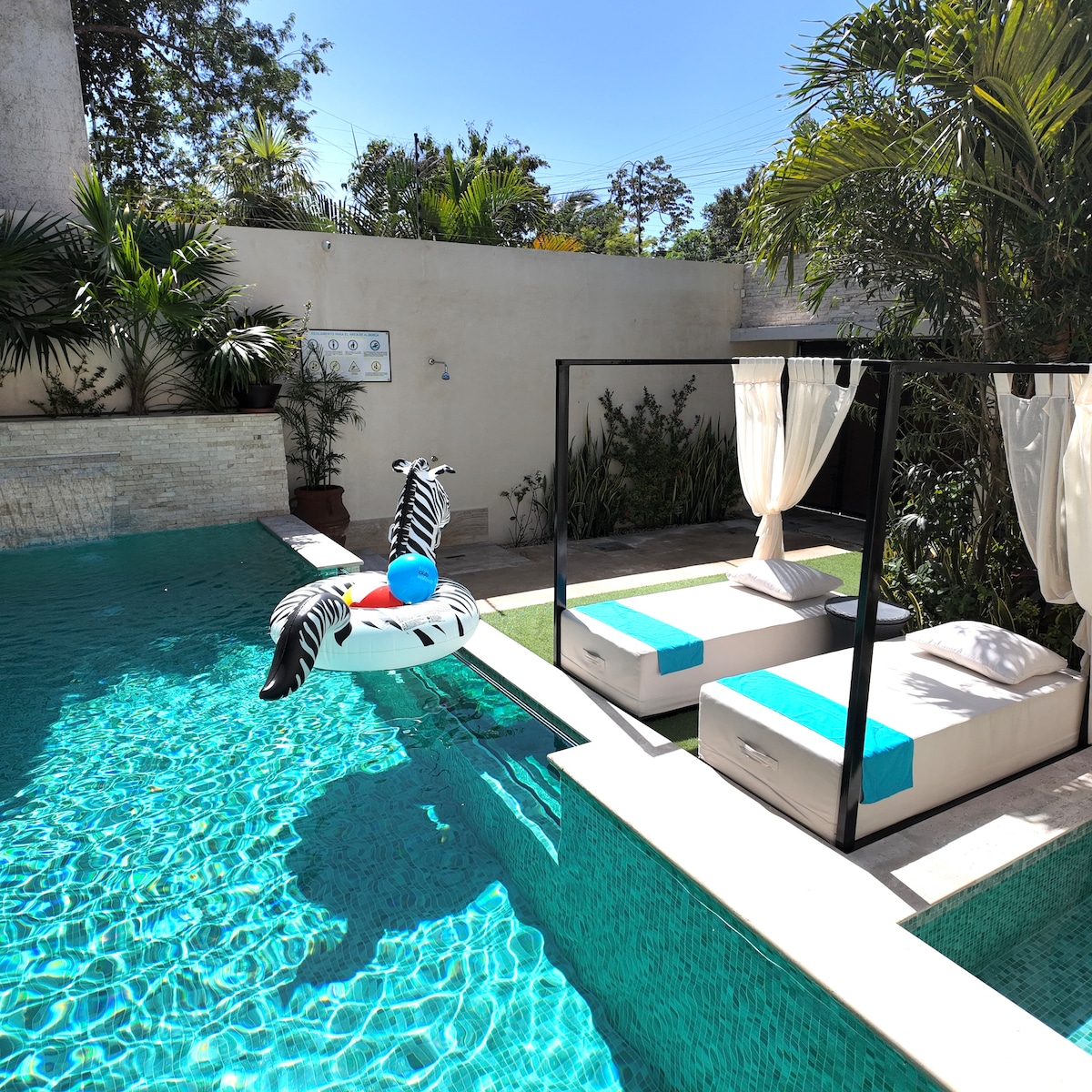 A vibrant outdoor pool area is showcased, featuring a large infinity pool surrounded by lush greenery. Two poolside loungers with turquoise cushions are positioned for relaxation. A playful inflatable float is seen drifting in the water, adding a touch of whimsy to the serene environment.