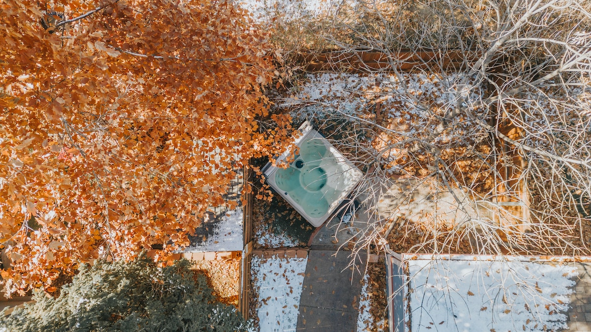 An outdoor space features a hot tub surrounded by autumn foliage, with a wooden pathway leading to it. The vibrant orange leaves contrast with the quieter colors of the nearby plants, creating a serene environment for relaxation.