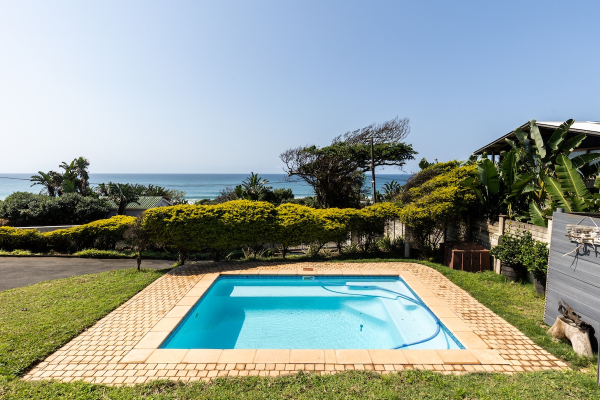 A small rectangular swimming pool is set within a tiled area, surrounded by a grassy lawn and lush greenery. The ocean can be seen in the background, adding a scenic view to the outdoor space.