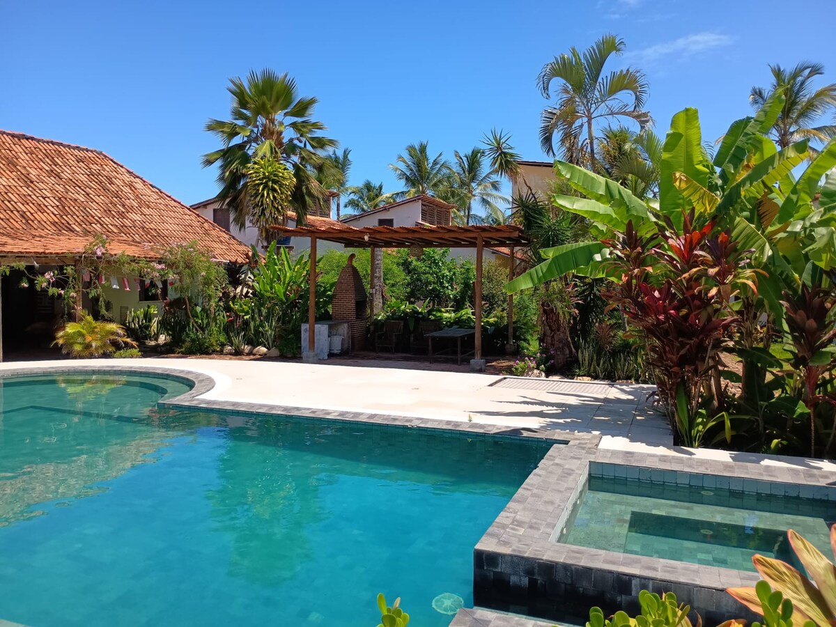 A serene outdoor space is presented, featuring a curved pool surrounded by lush tropical vegetation. A shaded area with wooden structure and seating is visible in the background, along with vibrant palm and banana plants adding to the tranquil atmosphere.