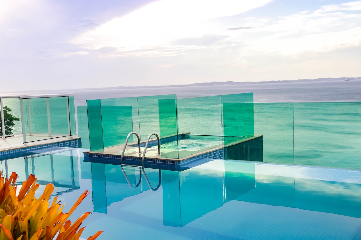 An expansive infinity pool offers unobstructed views of the ocean. Clear glass railings provide safety while maintaining the scenic vista. The water reflects the sky’s colors, creating a serene ambiance. Vibrant plants frame the pool, adding a touch of nature to the modern space.