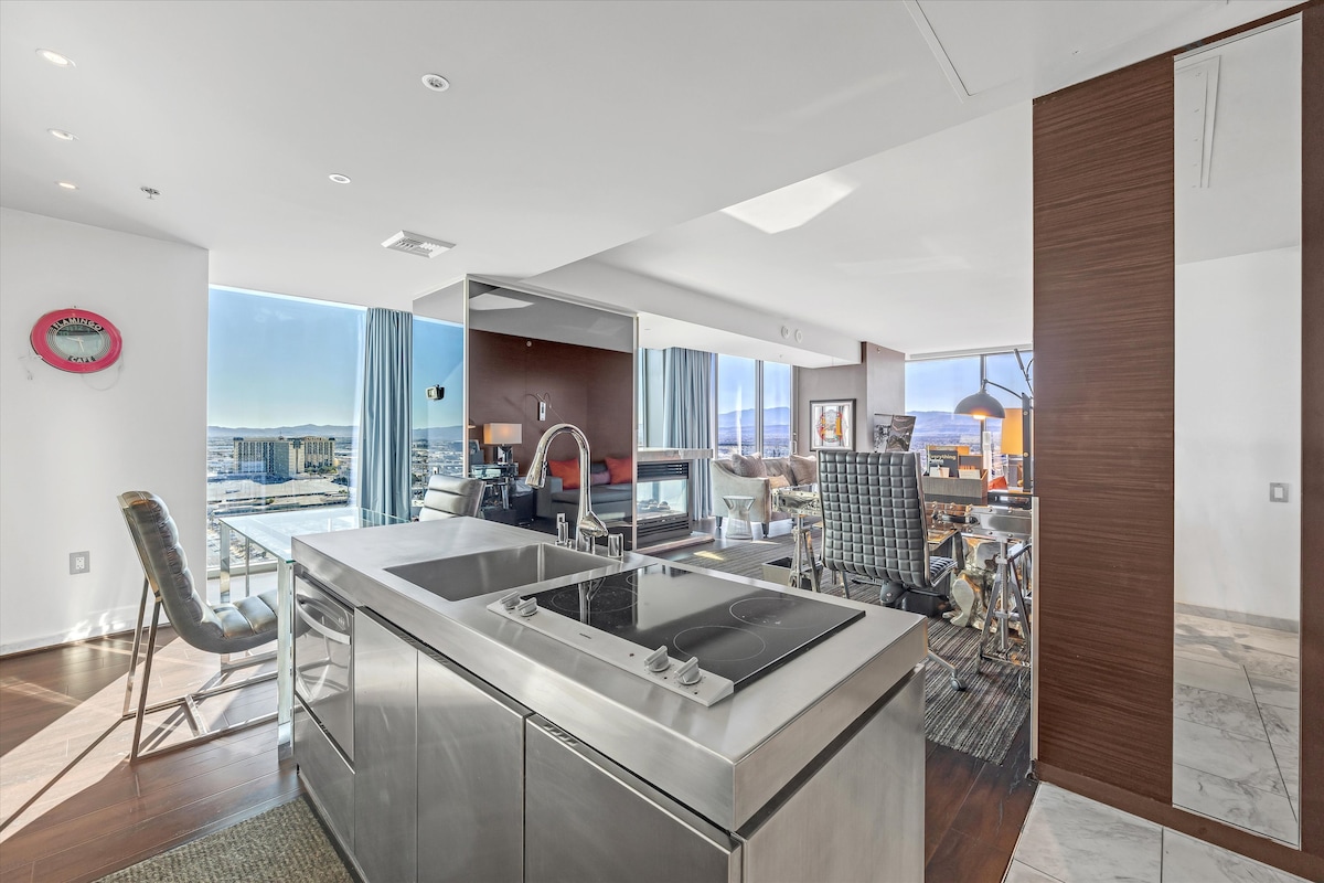 A modern kitchen area features sleek, stainless steel appliances and an island with a cooking surface. Large windows provide abundant natural light, offering views of the surrounding landscape. The open layout includes a dining area and comfortable seating, emphasizing spaciousness.