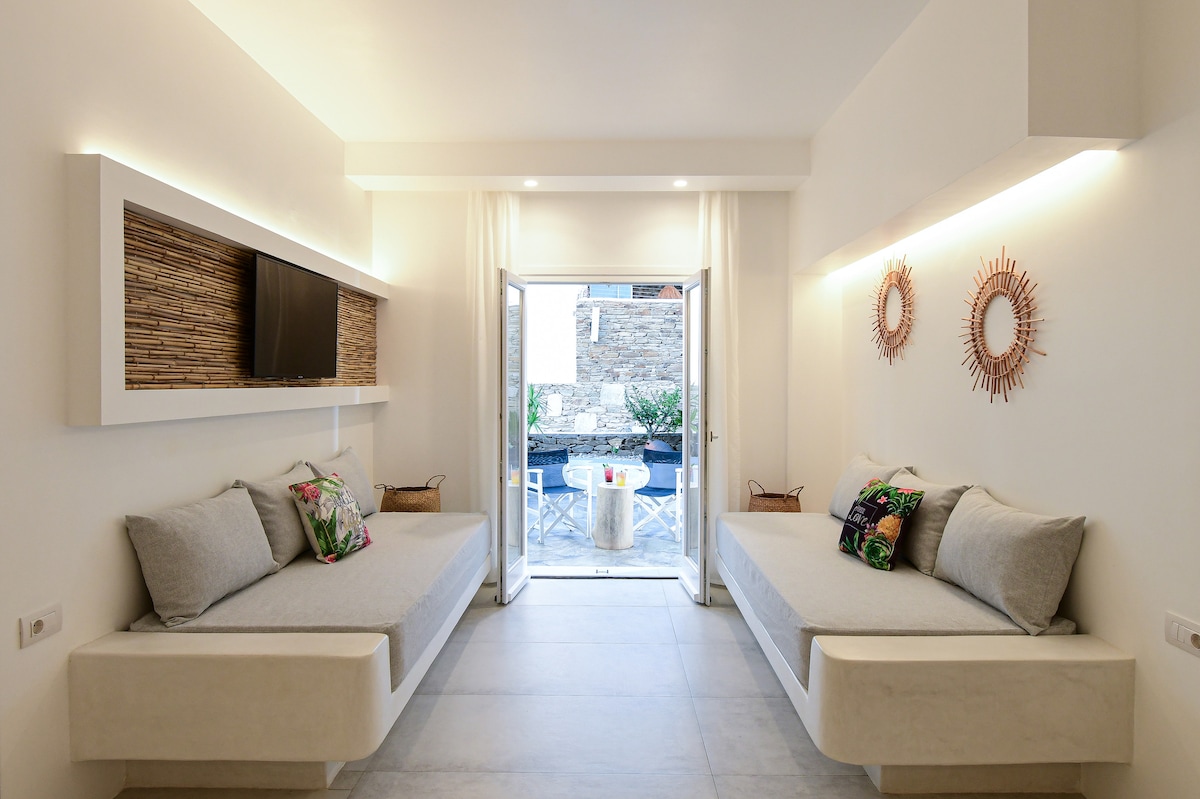 A bright sitting area features two spacious daybeds arranged against a light-colored wall. Natural textures are highlighted through woven decor. A large opening leads to an outdoor space, where a private pool is visible amid greenery.