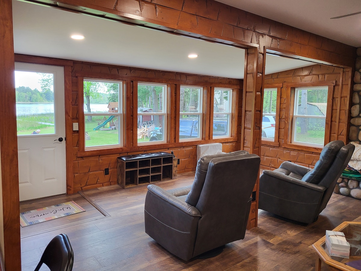 A spacious living area is highlighted by large windows that offer views of the surrounding nature. Seating is provided by two comfortable recliners facing a central area. The rustic wood finishes enhance the cabin's charm, with a welcoming entry door visible in the background.