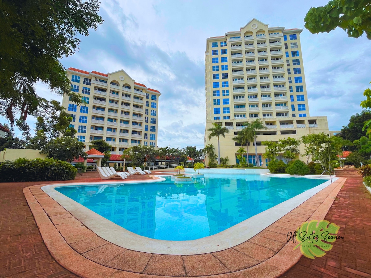 The image displays a sparkling pool surrounded by well-maintained gardens and lounge chairs. Two tall residential buildings are seen in the background, their modern architecture contributing to a spacious outdoor setting. Lush palm trees frame the area, enhancing the inviting atmosphere.