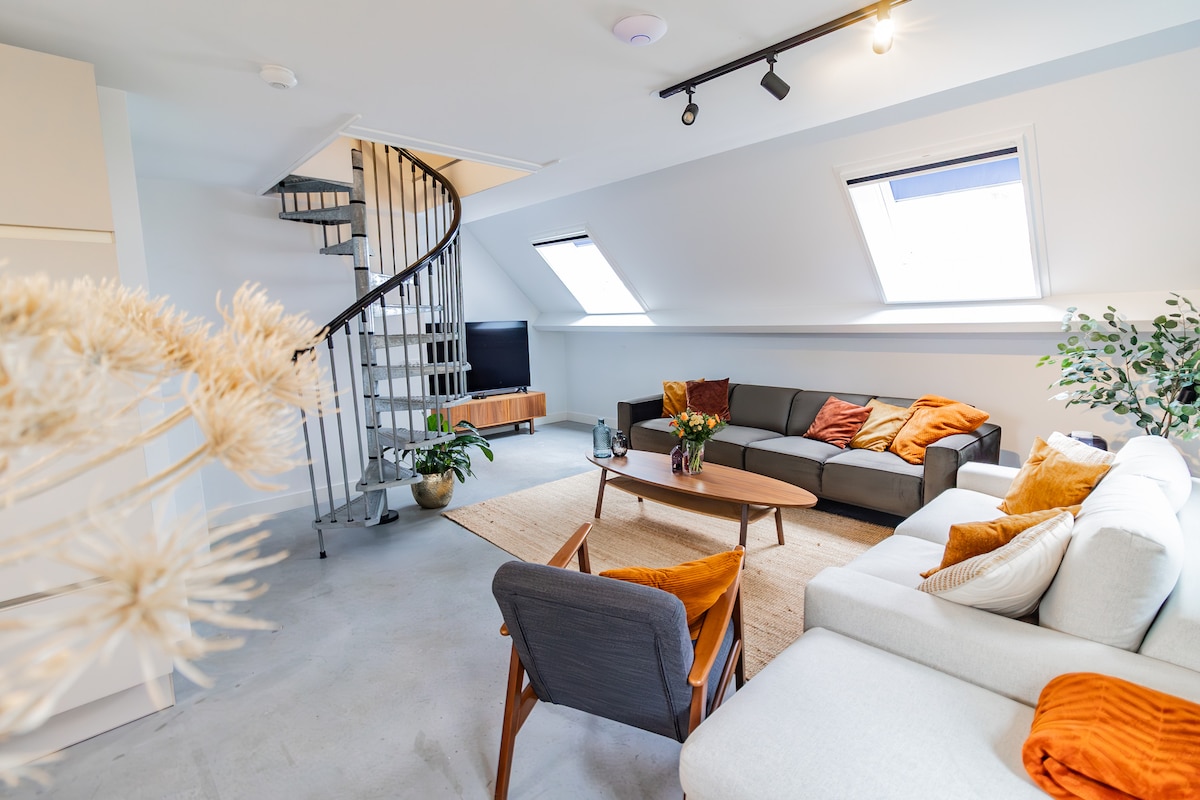 The spacious living area features a mix of seating options, including a light grey sectional sofa and a comfortable chair. Natural light enters through skylights, illuminating the room. A coffee table centers the space, adorned with flowers, while a spiral staircase provides access to the upper level.