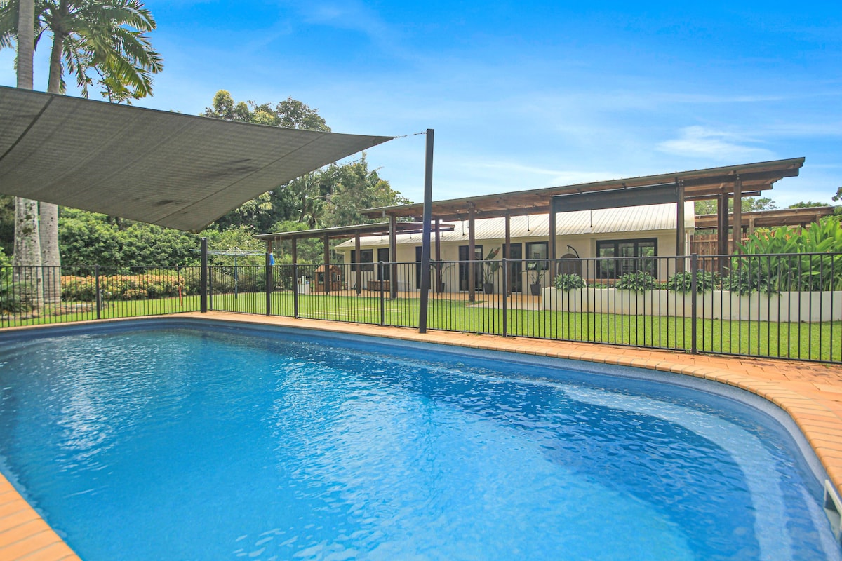 A clear view of a sparkling pool, surrounded by a protective fence and shaded by a large canopy. Lush greenery frames the background, with a modern single-story home featuring a spacious outdoor area visible beyond the pool.