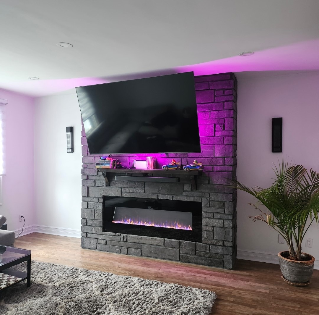 A modern living room features a large wall-mounted television above a stone fireplace. The fireplace is illuminated with a warm glow, and a green plant adds a touch of nature. Soft light filters through the window coverings, complementing the overall design.