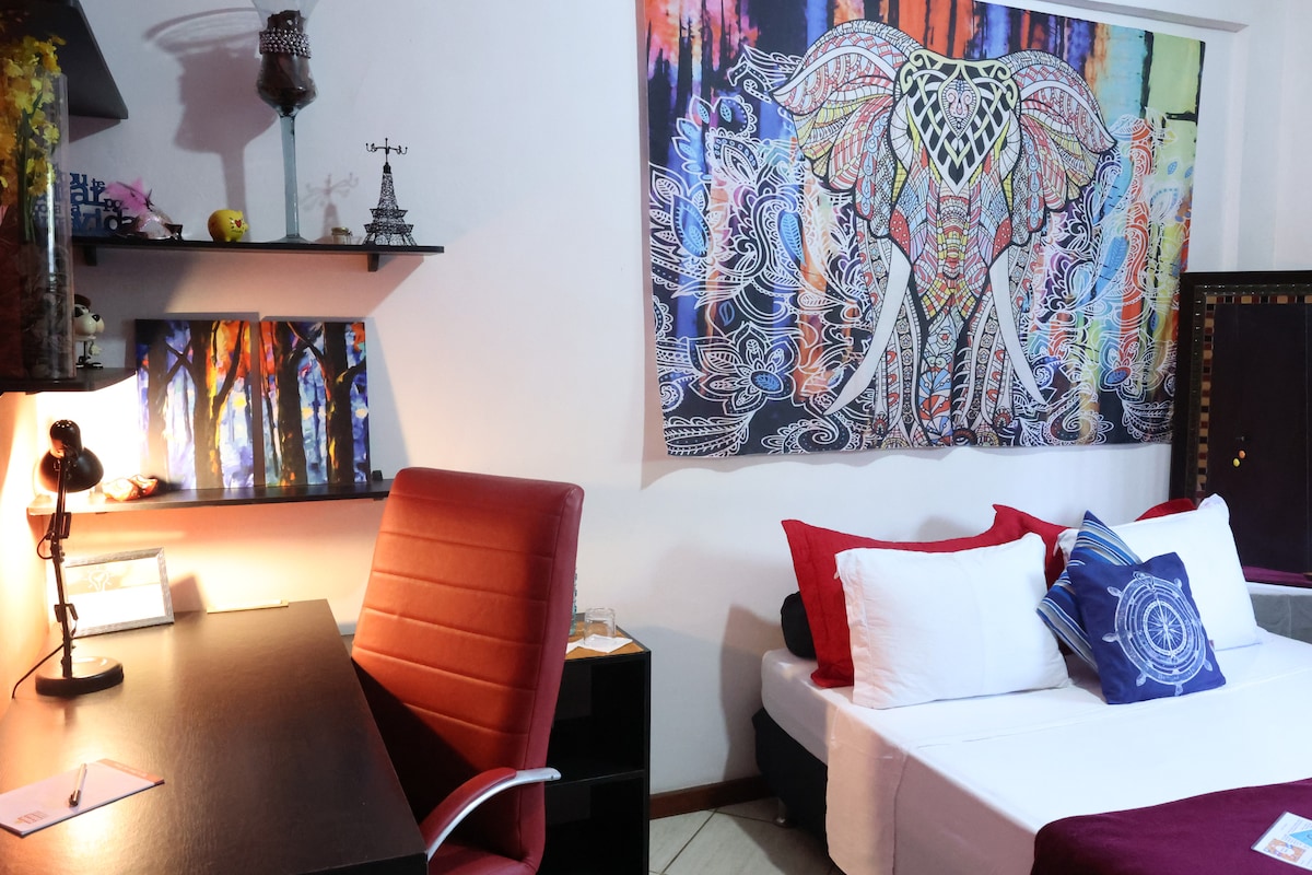 The room features a spacious bed adorned with white and blue pillows, complemented by a decorative elephant wall art. A desk with a lamp and chair is positioned nearby, offering an organized workspace. The colorful artwork on the wall adds vibrancy to the serene environment.