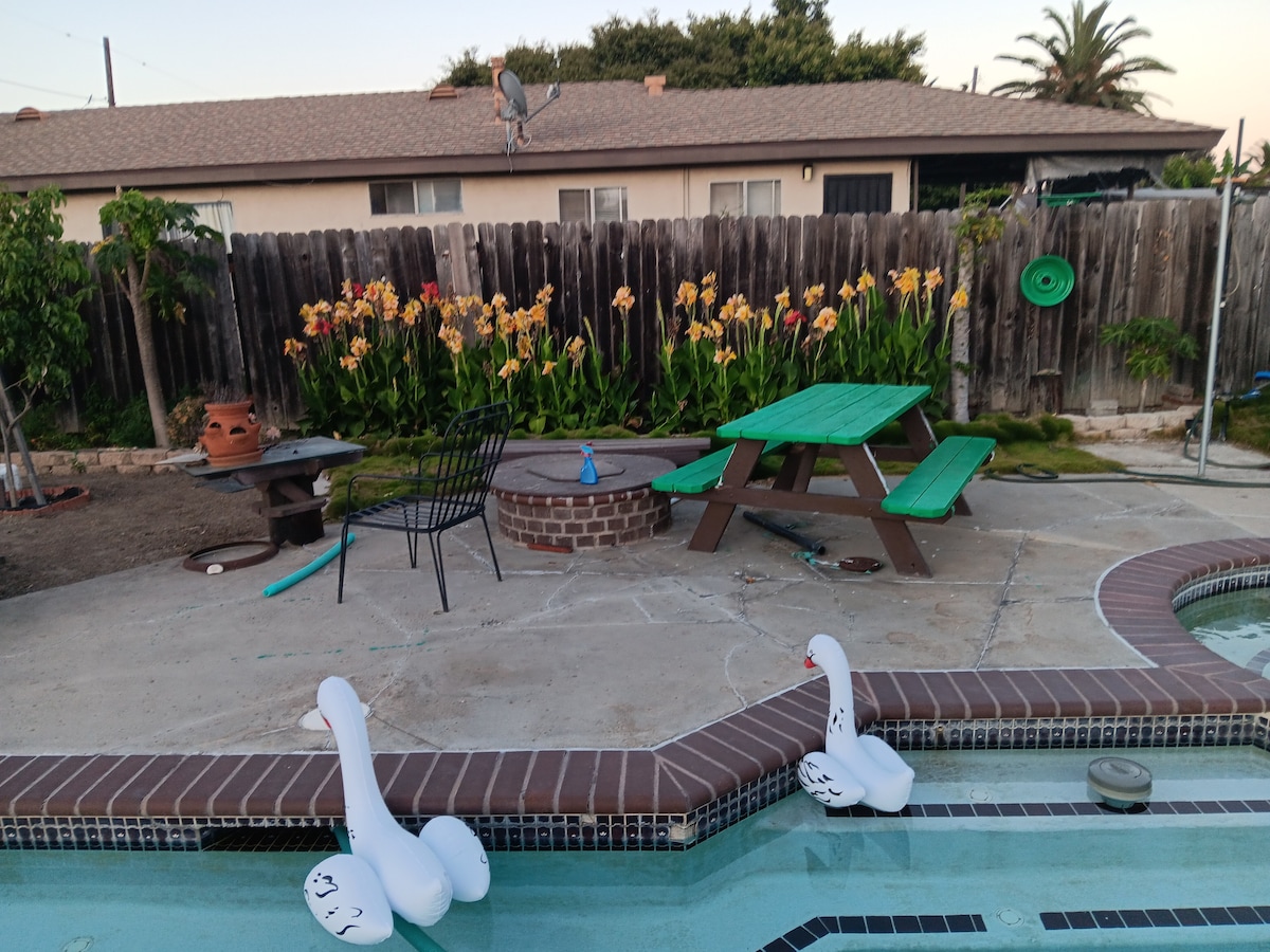 A private outdoor area features a lap pool with two white swan floaties. A circular seating area and a picnic table with vibrant green planks are visible. Flowering plants line the background, adding color to the space, while a wooden fence surrounds the property.