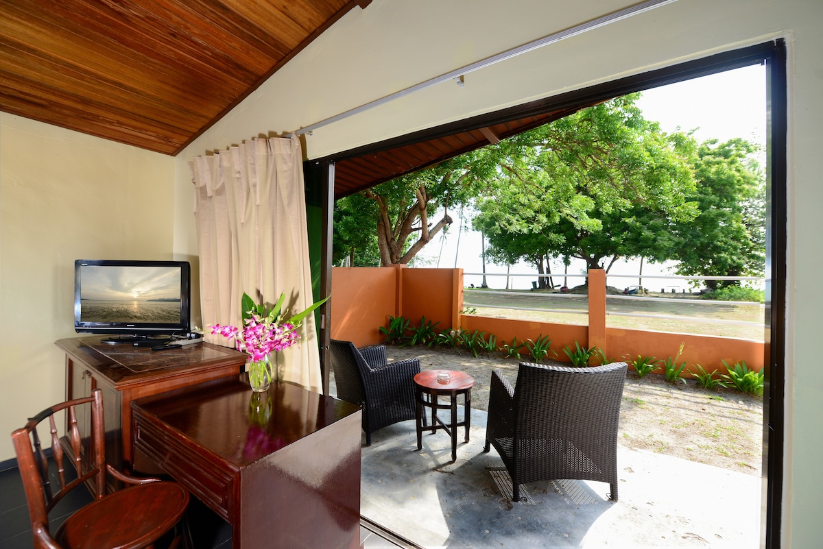 A cozy seating area is visible through the open sliding doors, featuring two wicker chairs and a small table. A wooden desk and television are positioned nearby, while lush greenery frames the view outside, enhancing the connection to nature.