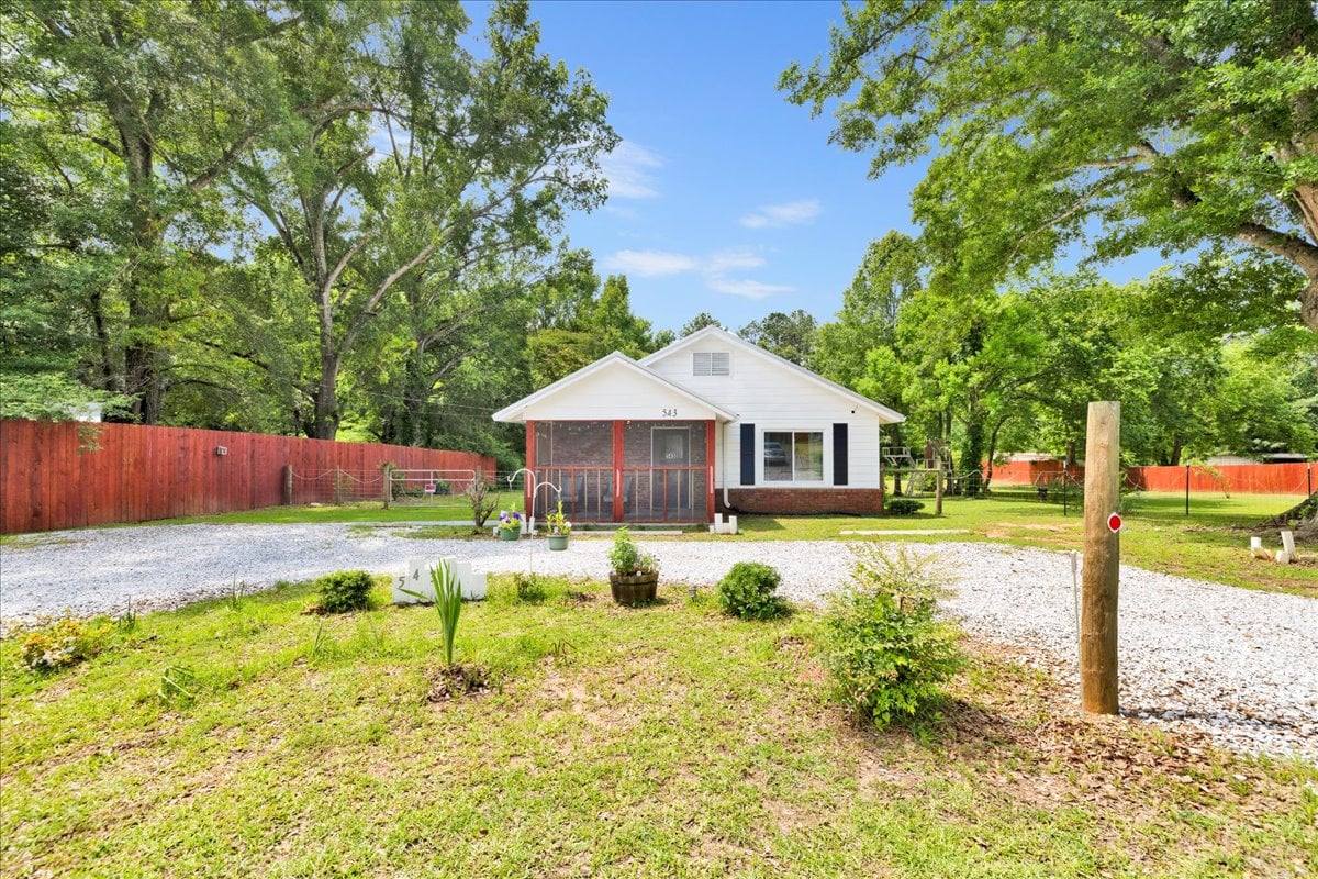 The exterior of the home is surrounded by a lush green lawn, bordered by mature trees. A gravel driveway leads to the front entrance, with flower beds and shrubs adding natural beauty. A privacy fence encloses the property, providing a sense of seclusion.