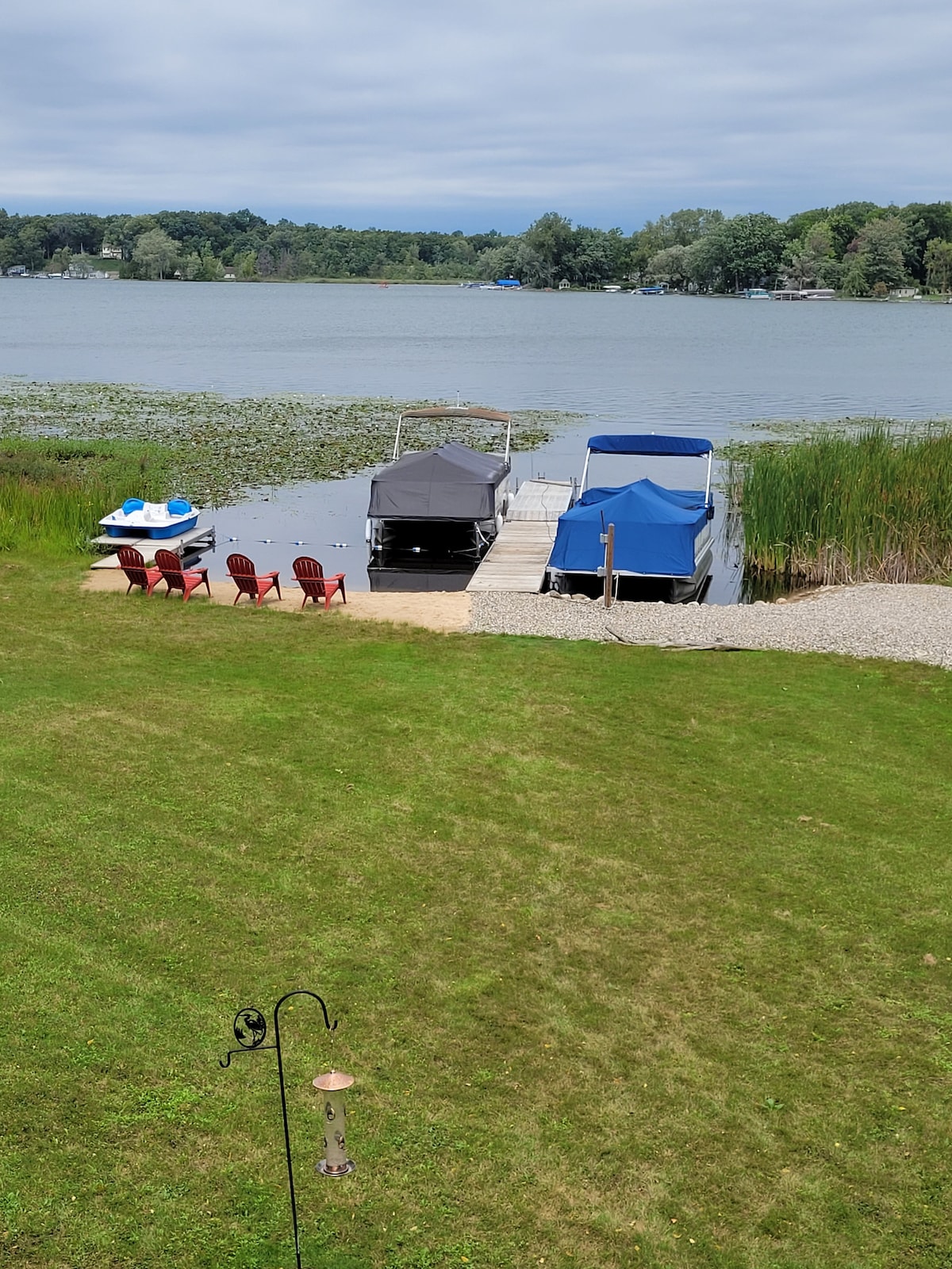 A private dock extends into the water, featuring two covered boat slips. Red outdoor chairs are arranged on the shore, providing a space for relaxation. Lush greenery and reeds frame the tranquil scene, with a view of the lake's surface reflecting the cloudy sky.