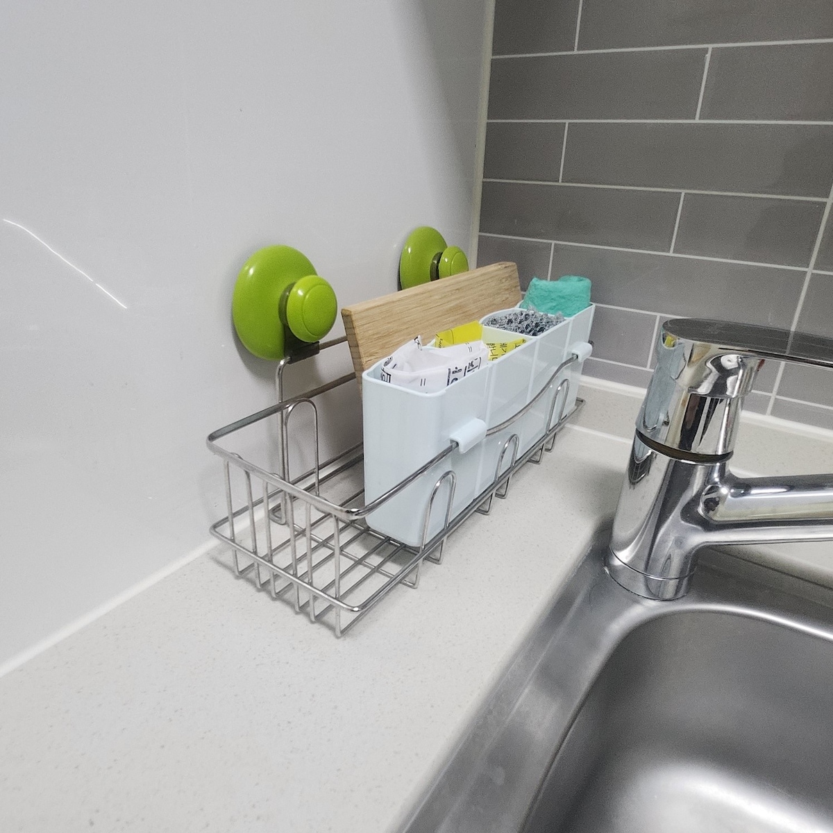 A kitchen sink area is featured, with a metallic rack holding utensils and sponges. Two bright green suction hooks are mounted on the wall, supporting wooden and colorful items. The sink is stainless steel, and a modern faucet is positioned alongside.