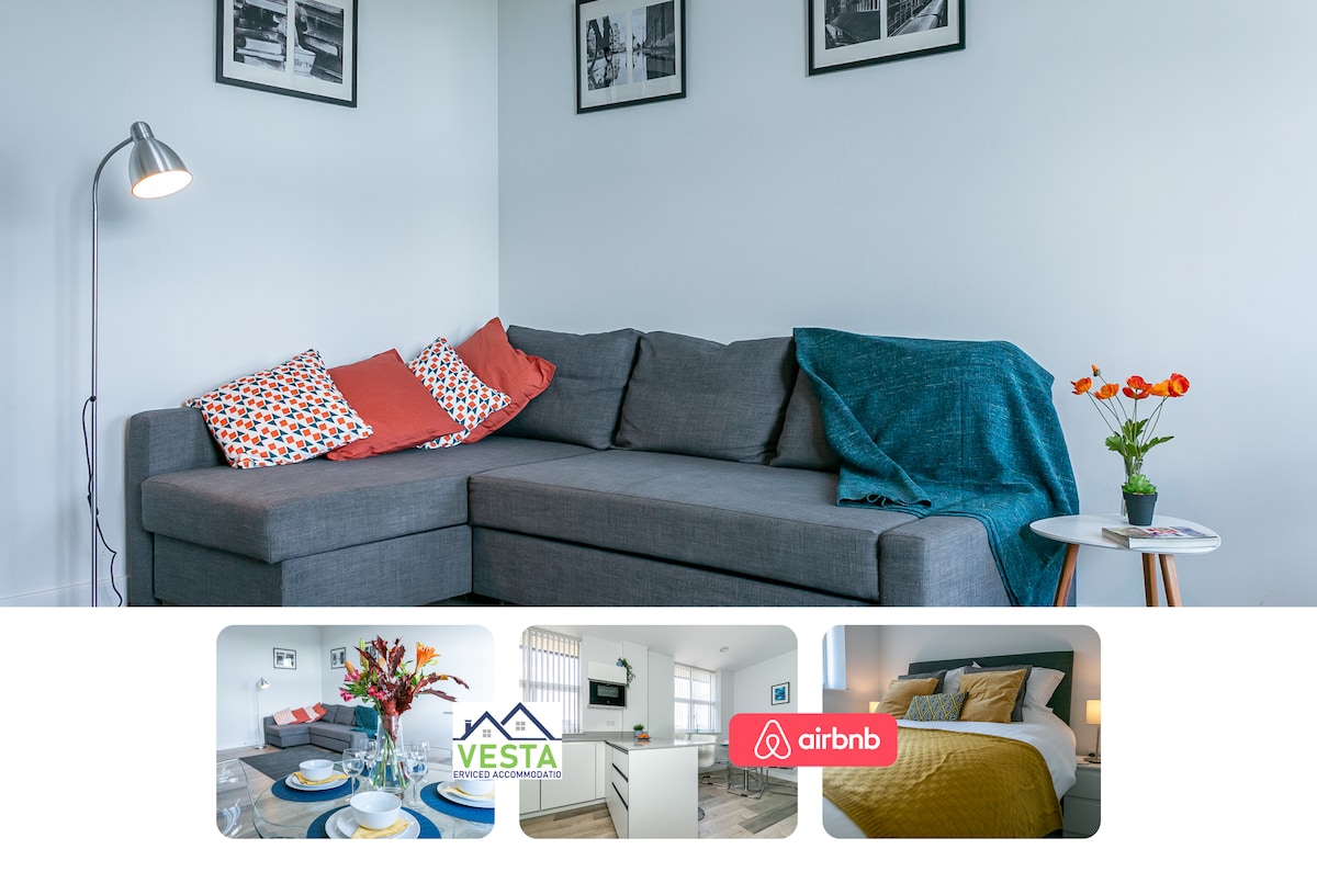 A spacious lounge area showcases a comfortable gray sectional sofa adorned with colorful cushions and a soft throw. A small round table is positioned nearby, featuring a vibrant bouquet. Light enters through a nearby window, creating a bright and welcoming environment.